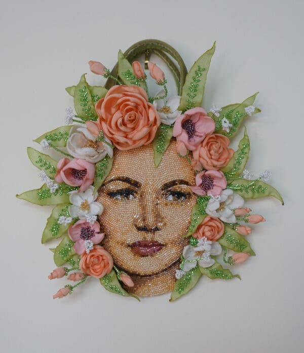 Decorative wall art featuring a mosaic face surrounded by a floral frame with pink and white flowers and green leaves, adorned with glitter.